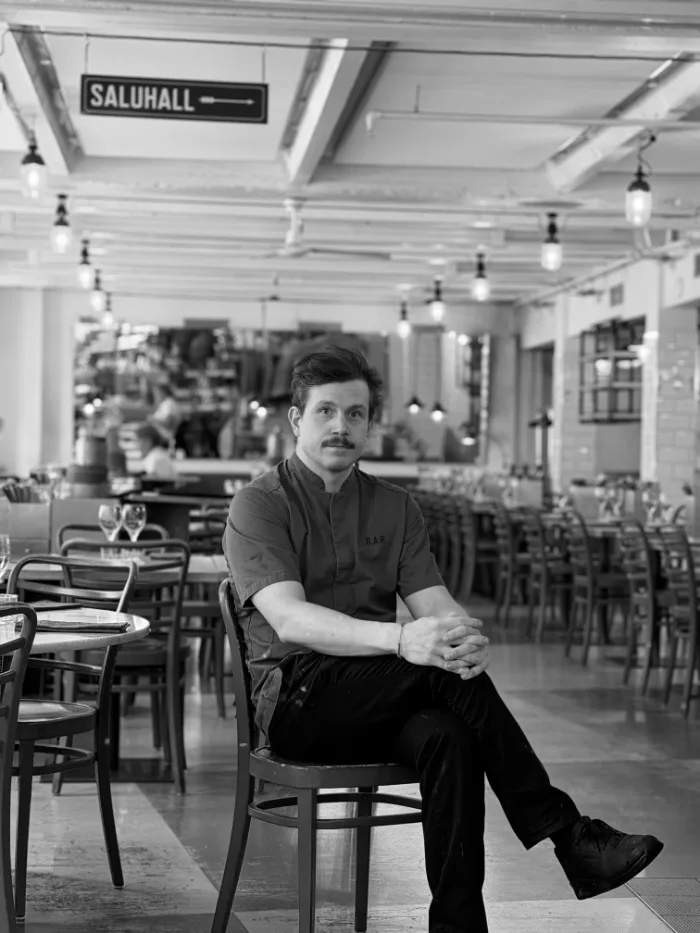 Medarbetare på B.A.R. i mörk skjorta och mustasch, händerna i knät, sittande i matsalen under skylten Saluhall