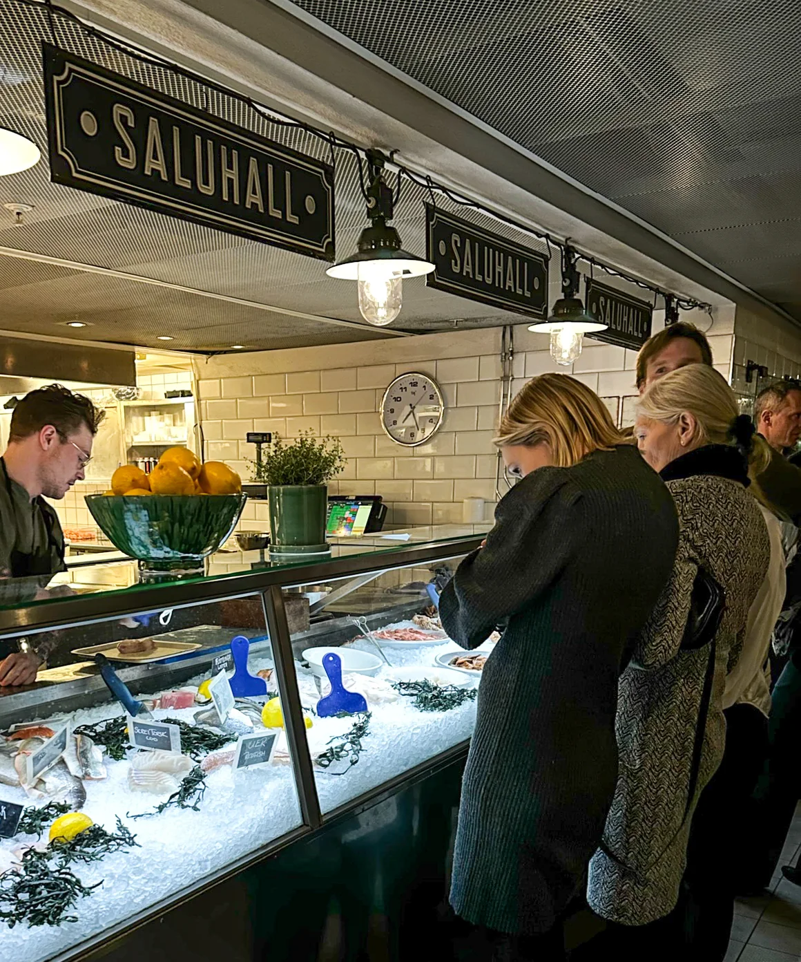 Gäster vid ismontern under skyltarna med texten Saluhall, med färsk fisk på is bakom glaset