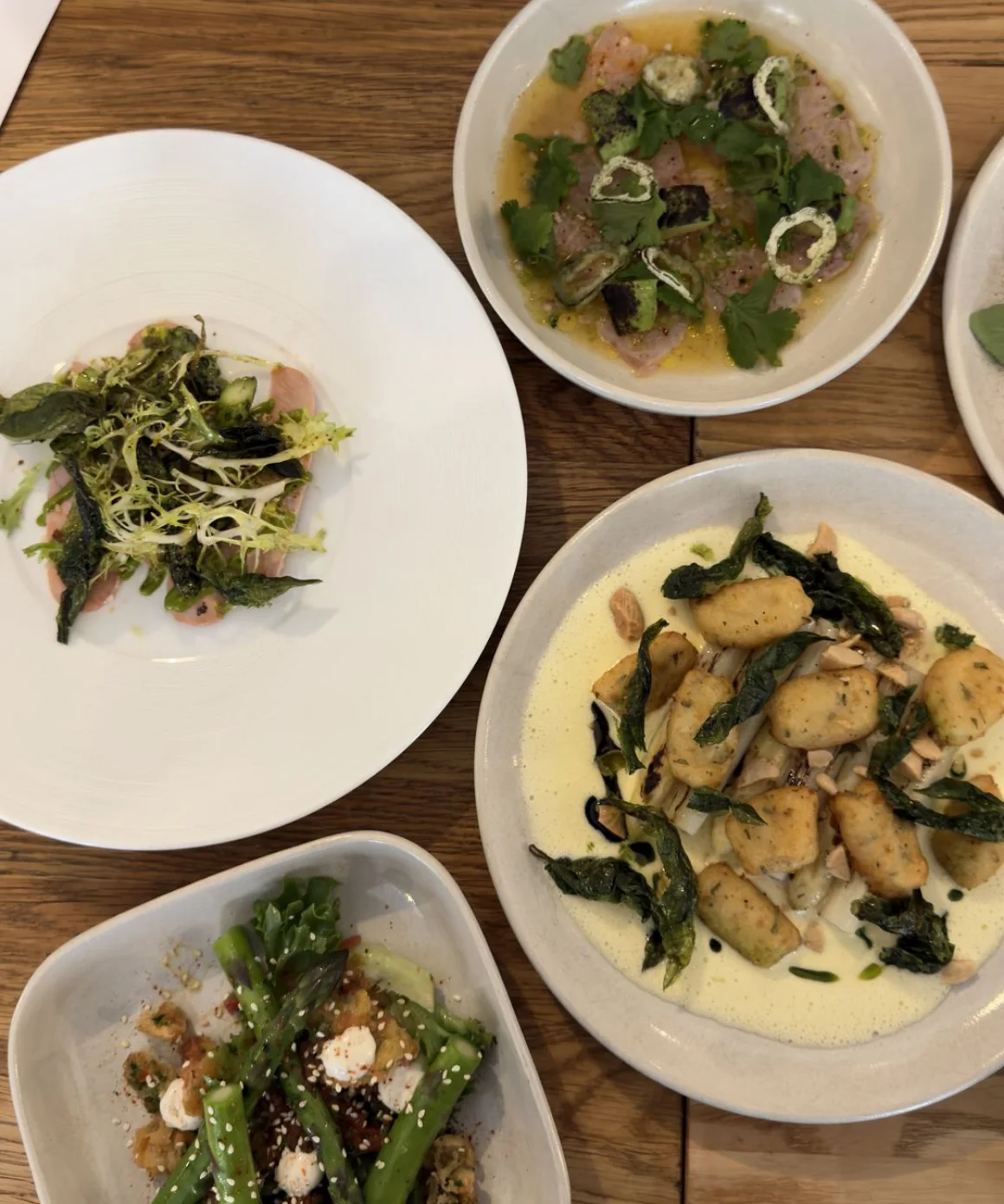 Flera tallrikar på ett träbord: tunna fiskskivor under frisésallad och rostade blad, en skål med fisk i gul buljong med koriander och jalapeño, och gnocchi i ljus sås med friterade blad